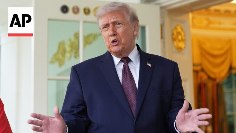 Man in a navy suit and red tie speaks with open hands outside a doorway, AP logo visible on the left.