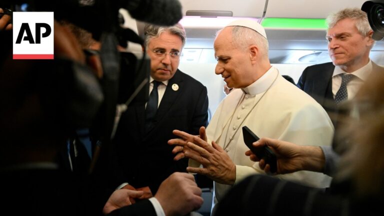 Pope Francis on a plane, in white robes, surrounded by reporters with phones and cameras; AP logo visible left.