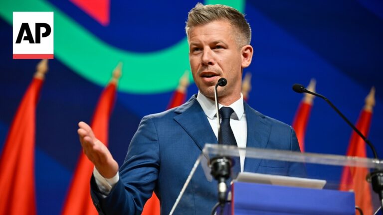 Politician in a blue suit speaks at a podium with microphones, AP logo in the corner and red flags in the background.