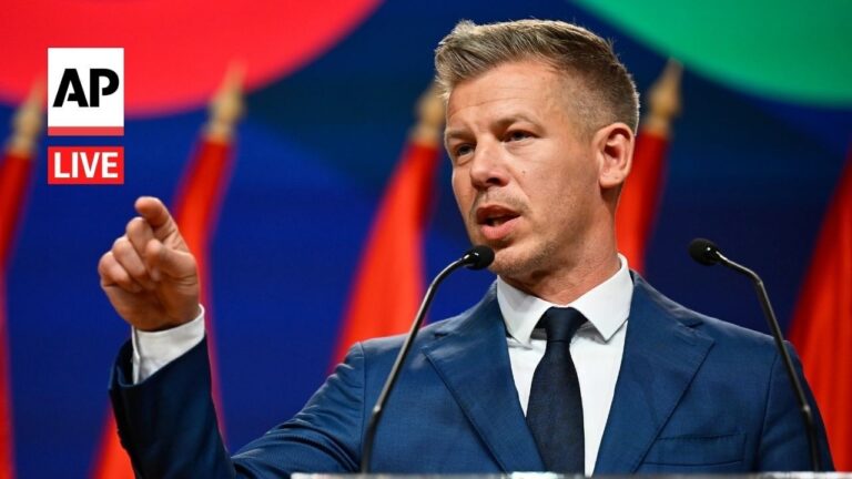 Man in a blue suit speaks at a podium, pointing with his right hand, AP Live logo in the upper left corner behind red flags behind him.