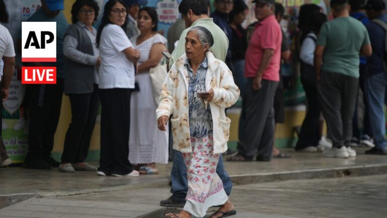 Older woman in a floral dress and white fleece jacket walks on a sidewalk, holding a phone, with a crowd behind her.