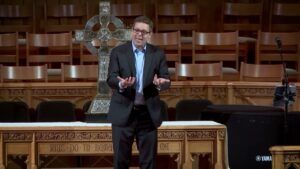 Man in a dark suit giving a sermon on a wooden pulpit, with a large metal cross behind him.