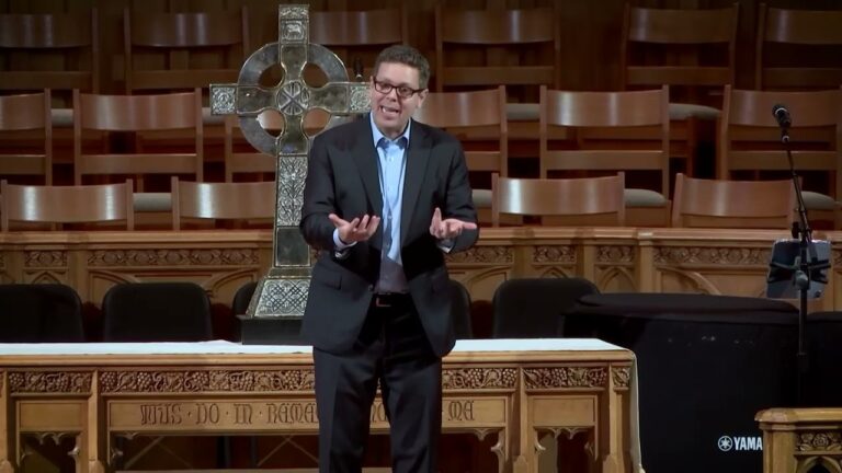 Man in a dark suit giving a sermon on a wooden pulpit, with a large metal cross behind him.
