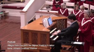 Organist in a dark suit plays a wooden pipe organ as a choir in red robes looks on in a church; on-screen text shows a classical piece title and composer.