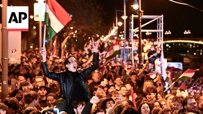 Nighttime crowd at a rally waving Hungarian flags; a man in a black leather jacket is lifted on someone’s shoulders, arms raised and cheering, AP watermark in the top-left.