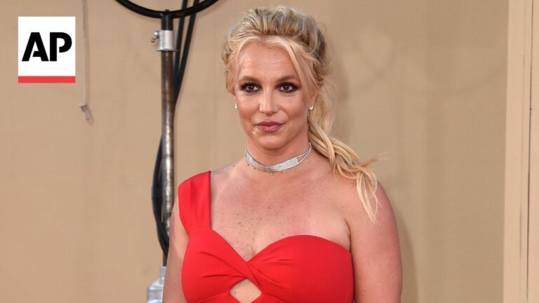 Blonde woman in a red one-shoulder dress posing on a red carpet, AP logo in the upper left corner