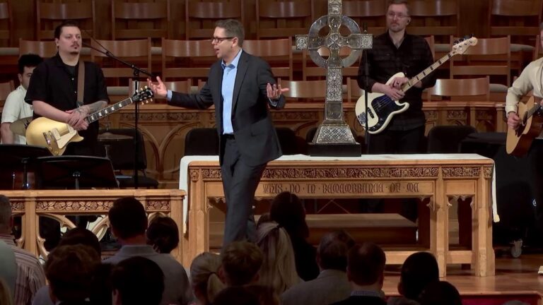 Speaker in a suit addressing a worship band on a wooden stage with a large ornate cross and altar behind him; audience seated in front.