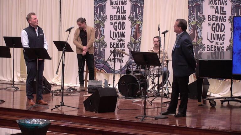 Stage with five men on a church-like platform; speaker in a vest at left, others with microphones, drummer behind, and a man in a suit at right; banners behind read 'Trusting that all belong to God living'.