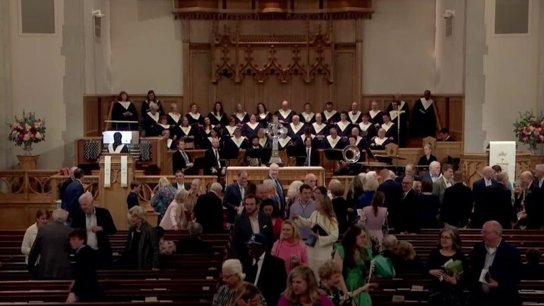 Choir in black robes on risers behind an ornate wooden organ while people mingle in the pews of a large church.
