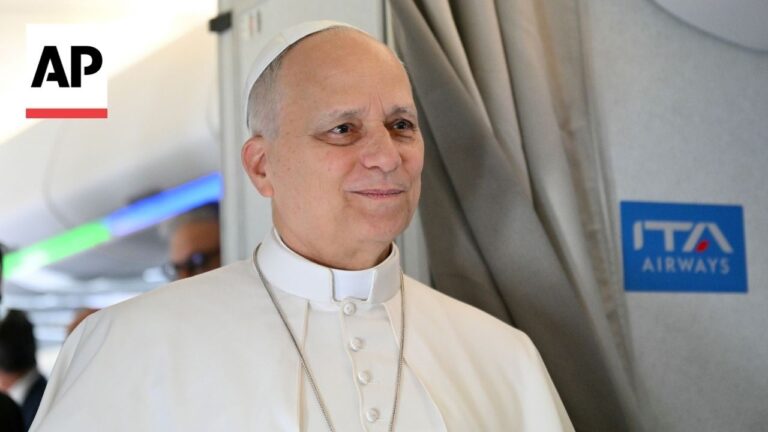Pope Francis in white cassock smiling on an airplane, AP News logo in the top-left corner and an ITA Airways sign in the background.