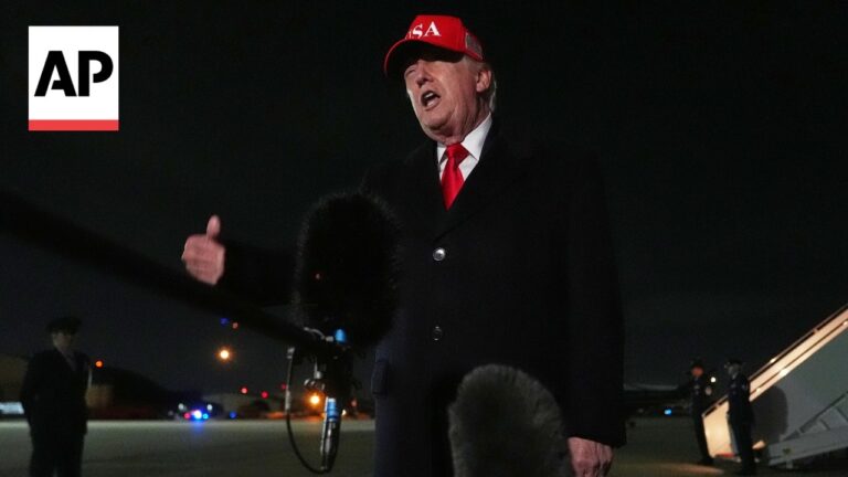 Male political figure wearing a red USA cap speaks into multiple microphones on a dim tarmac at night, AP logo in corner.
