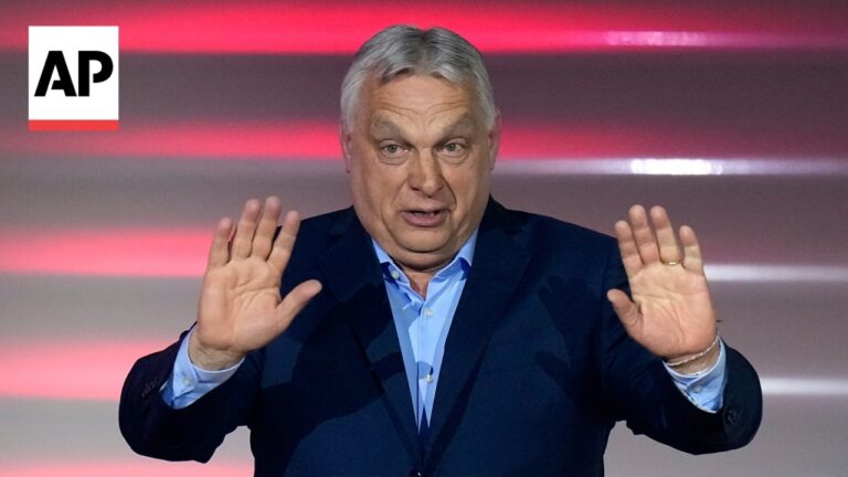 Man in a dark suit raises both hands on stage, pink-lit background, AP logo in the top-left corner.