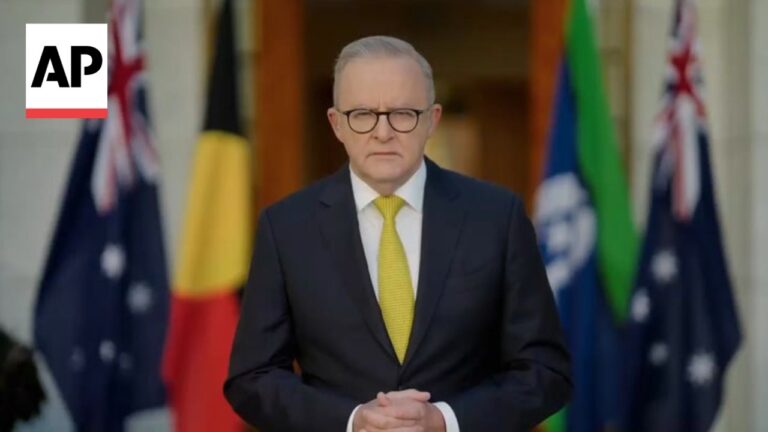 Man in a dark suit and yellow tie stands with hands folded in front, flags behind him, AP logo in the top-left corner.