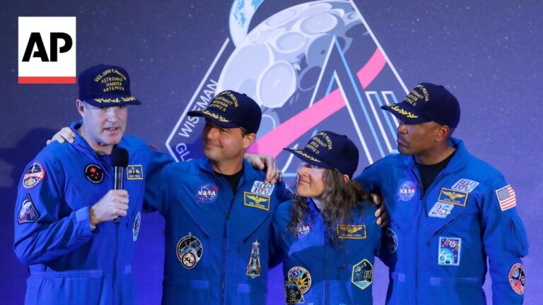 AP logo at left; four astronauts in blue jumpsuits and caps stand together on a space-themed stage, one holding a microphone for an interview.