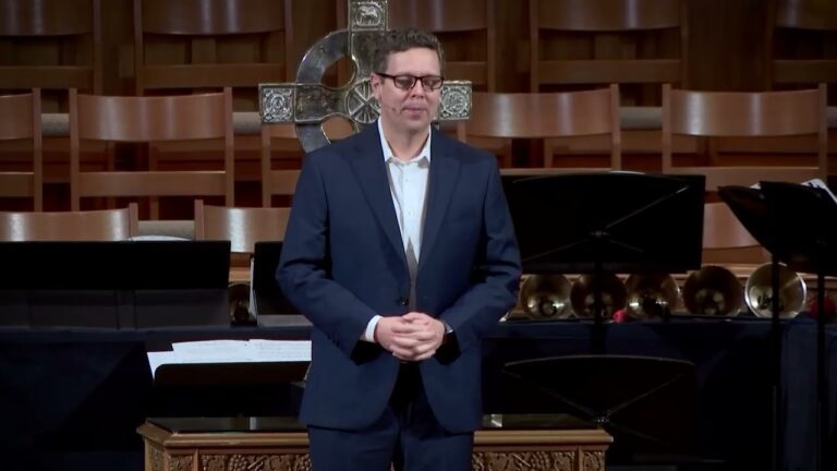Man in a navy suit and glasses stands at a lectern in a church, hands folded, with a large decorative cross behind him.