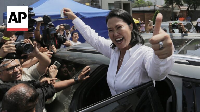 Smiling woman in a white blouse gives a thumbs up from a car window as reporters with cameras surround her,