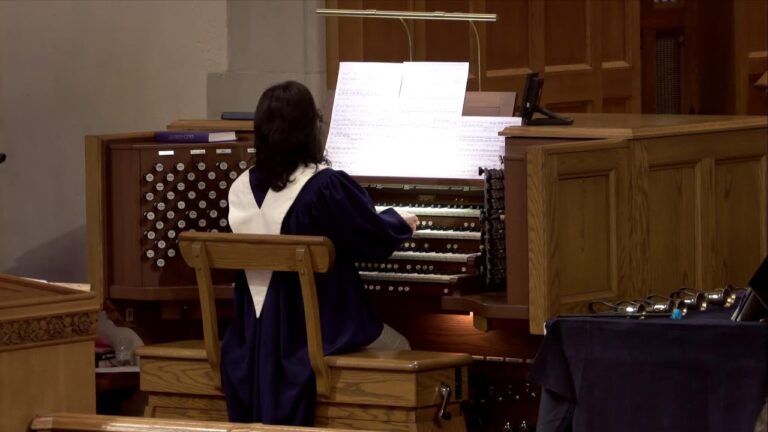 Person in a dark choir robe seated at a multi-keyboard pipe organ, reading sheet music in a church setting.