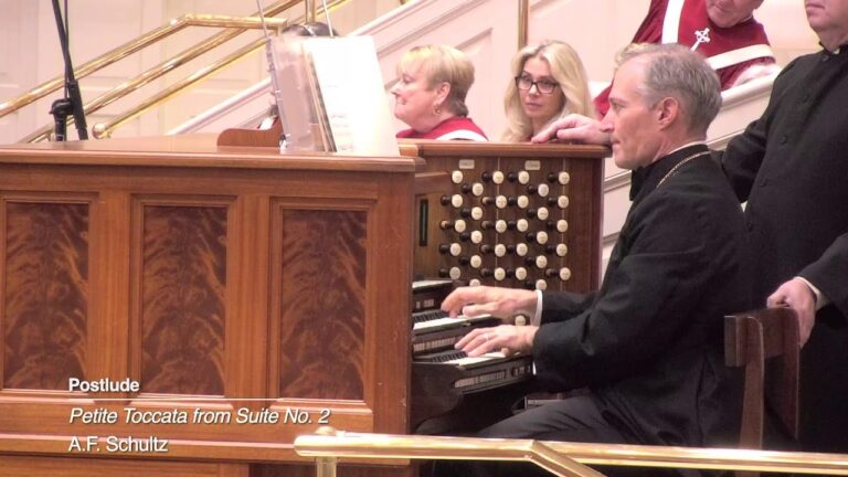 Organist in black robes plays a wooden pipe organ as choir members look on.