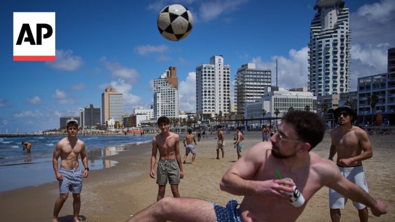 Men play beach soccer on a sunny day, ball in midair near the shore, with a city skyline and AP logo in the top-left.