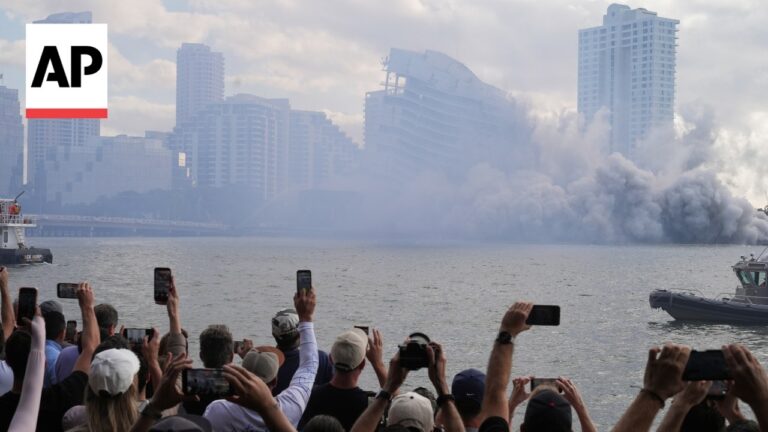 Crowd of onlookers taking pictures of a heavily damaged, collapsing high-rise with smoke rising over a riverfront cityscape. AP logo visible in the top left.