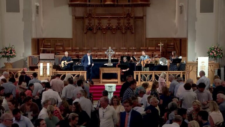 Church sanctuary with a live band on stage; guitarist, keyboard, and vocalist perform as the congregation mingles in the foreground.