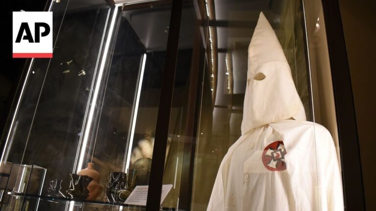 White hooded robe on display in a glass case, with a red circular chest patch bearing a dark insignia; AP watermark in the corner.