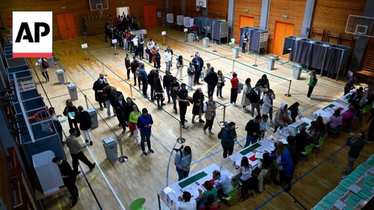 Indoor gymnasium used as a polling station with a long line of voters waiting, ballot booths along the walls, and tables with officials assisting voters; AP logo watermark in the top-left corner.