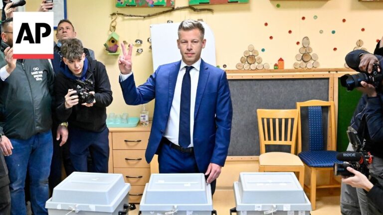 Man in a blue suit posing with a peace sign beside three ballot boxes while photographers film him in a classroom or polling-site setting with an AP logo overlay on the left.