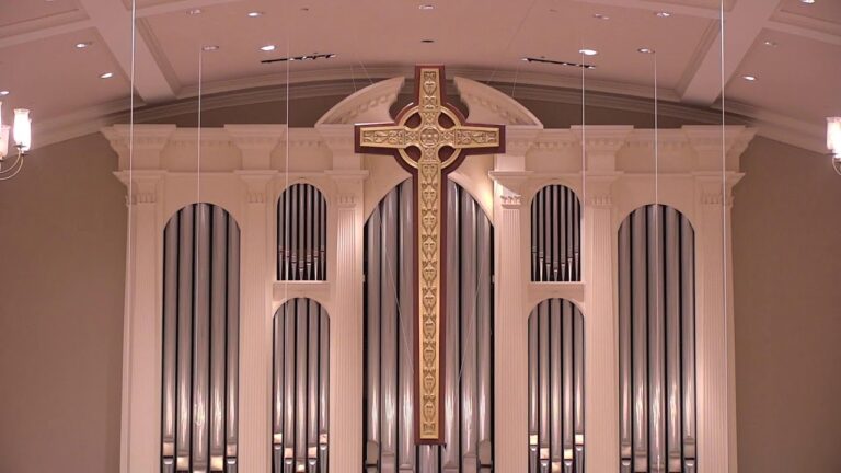 Interior of a church sanctuary featuring a large decorative cross hanging in front of a pipe organ backdrop.