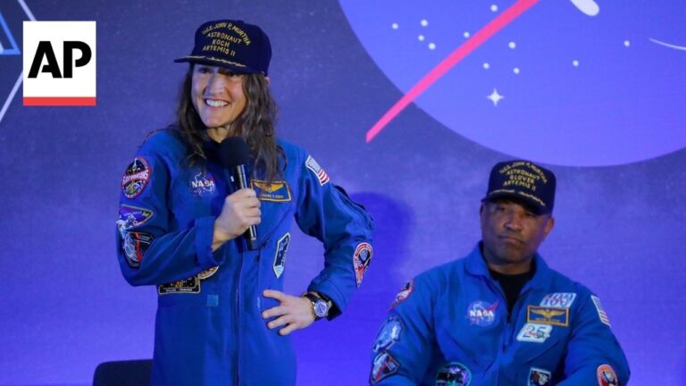 Two astronauts in blue NASA-style jumpsuits on a stage; a smiling woman stands with a microphone in hand while a man sits beside her, AP logo on the left.