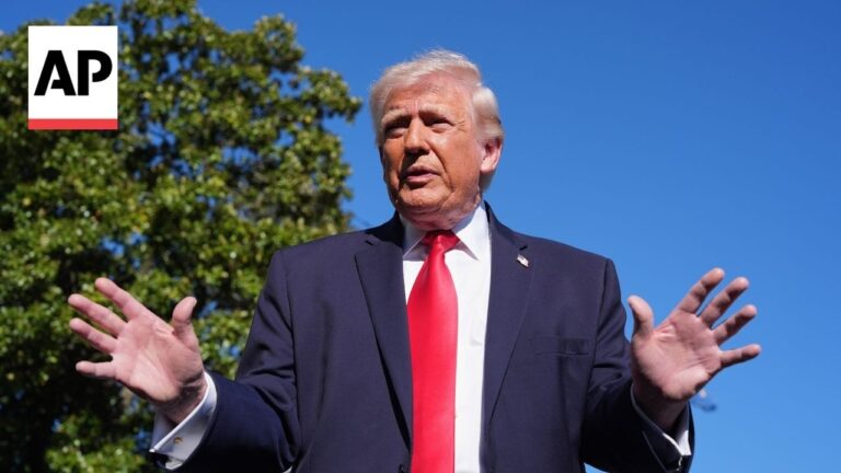Man in a navy suit with a red tie speaks outdoors, hands raised; blue sky and trees in the background, AP logo in the corner.