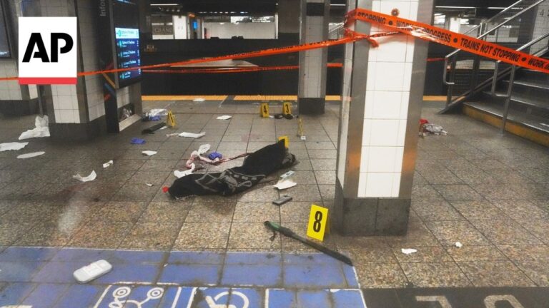 Subway platform cordoned off with orange tape and yellow evidence markers; scattered papers, clothing, and debris on the tiled floor.