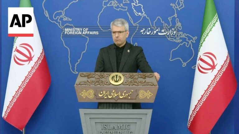 Iranian official at a press conference podium with flags on both sides and a world map backdrop, AP watermark in corner.