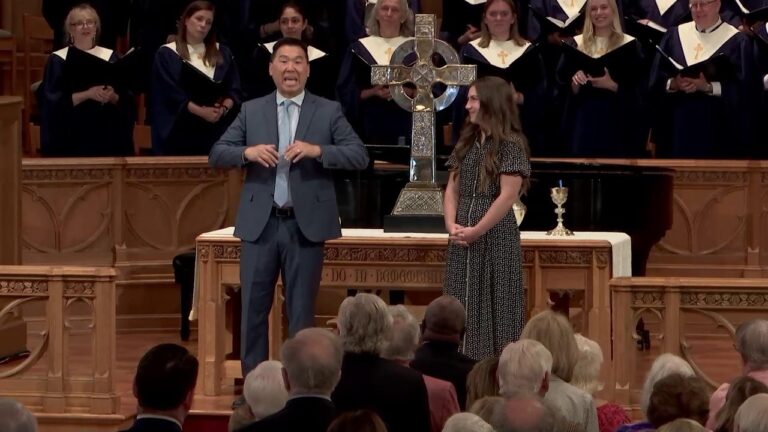 Man in a blue suit speaks at a church altar beside a standing woman, with a choir in robes behind them.