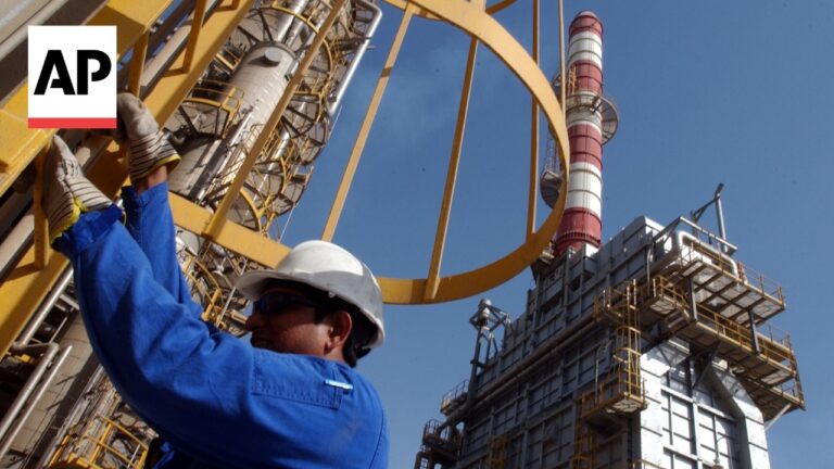 Industrial worker in blue coveralls and white hard hat adjusts yellow steel framework at an oil refinery scene, AP watermark visible in corner