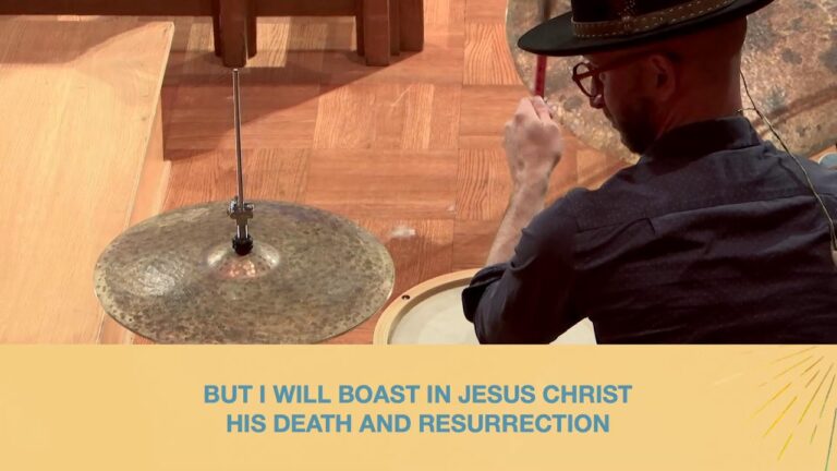 Drummer in a hat and glasses plays a cymbal on a wooden stage; a yellow banner at the bottom reads, “But I will boast in Jesus Christ His death and resurrection.”