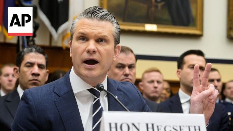 Man in a blue suit testifying at a hearing, speaking into a microphone and showing a three-finger gesture; AP logo visible in corner.