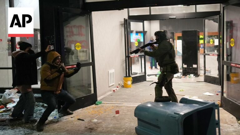 Armed officer in tactical gear aiming a gun at two civilians near a store entrance with shattered glass and debris outside, AP watermark top left.