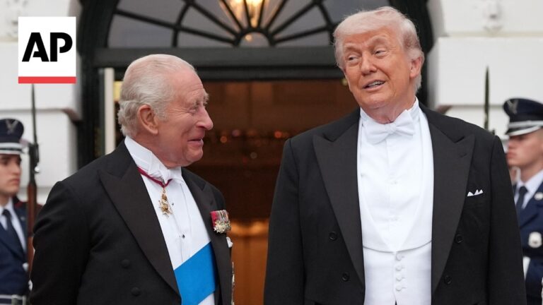 Two men in black tuxedos with bow ties smile and chat outside a grand doorway; one wears a blue sash and medals, AP logo in the corner.