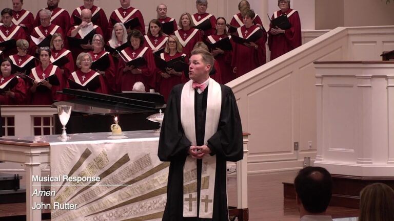 Male conductor in a black robe with a white stole leads a burgundy-clad choir from a pulpit in a church setting, with a grand piano behind.