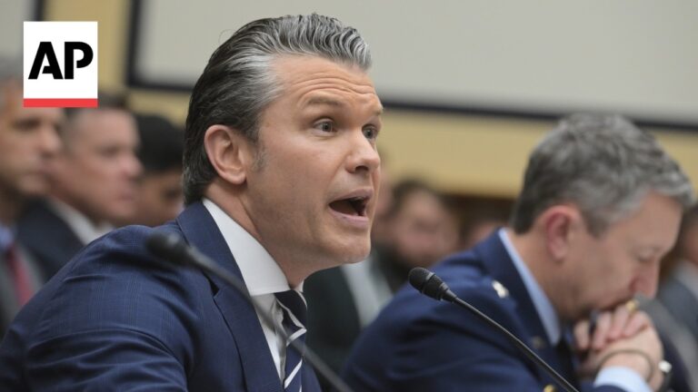Man in a navy suit speaks into a microphone at a formal hearing, AP logo visible in the top-left corner.