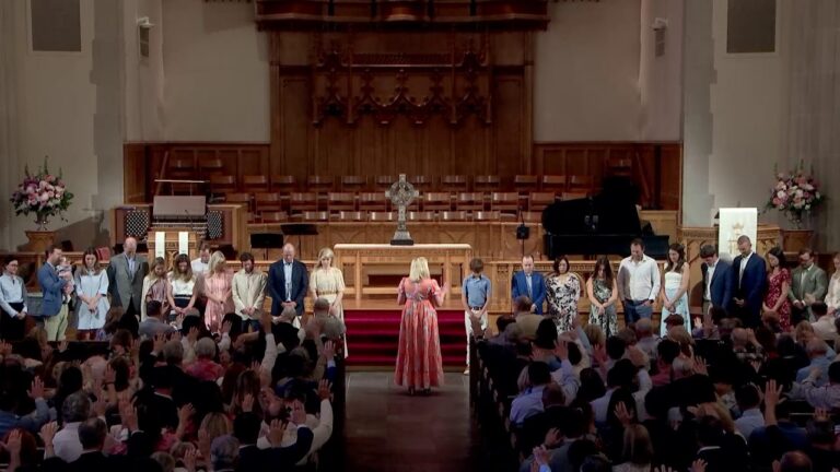 A woman in a coral dress conducts a line of people on a church stage as the audience raises hands in a ceremony.