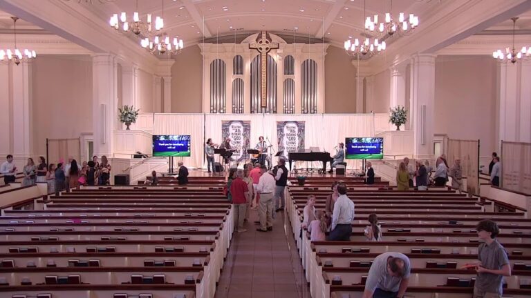 Interior of a church sanctuary with a live band performing on stage near a large cross and organ pipes, while attendees mingle in the pews.