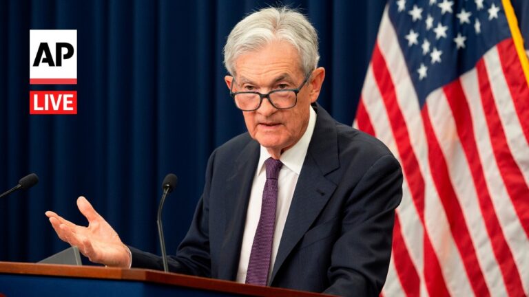Man in a suit and glasses speaks at a podium during a press conference, with the US flag in the background and an AP Live logo to the left.