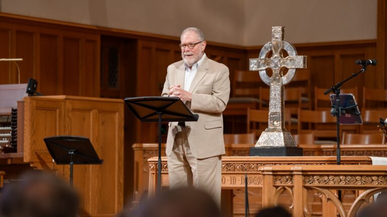 Older man in a beige suit speaks at a lectern on a wooden church stage, with a large metal cross on the altar behind him.