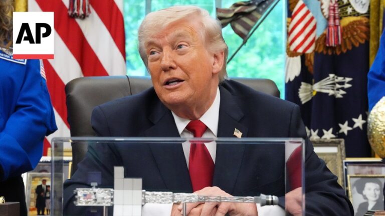 Man in a dark suit and red tie sits at a desk with a clear protective barrier, AP logo in the corner, American flag and other flags in the background.