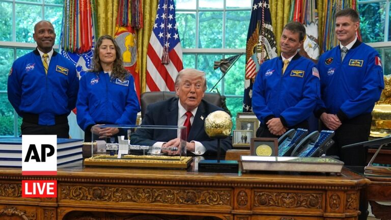 President Trump sits at an ornate desk in the Oval Office, flanked by astronauts in blue NASA jackets; AP Live watermark bottom left.