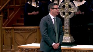 Man in teal suit and green tie speaks into a headset mic at a church lectern, with a large silver cross and choir behind him.