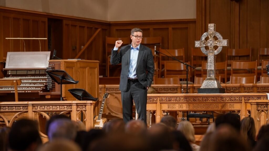 A man in a suit delivering a talk from a wooden pulpit in a church, with a decorative cross on the altar and an audience in front.