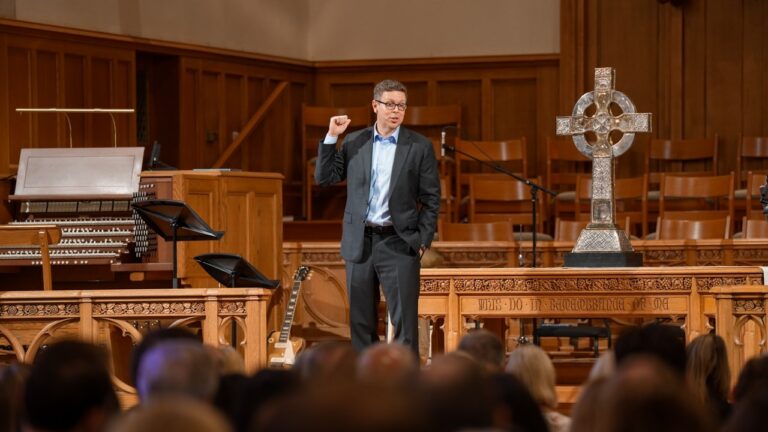 A man in a suit delivering a talk from a wooden pulpit in a church, with a decorative cross on the altar and an audience in front.